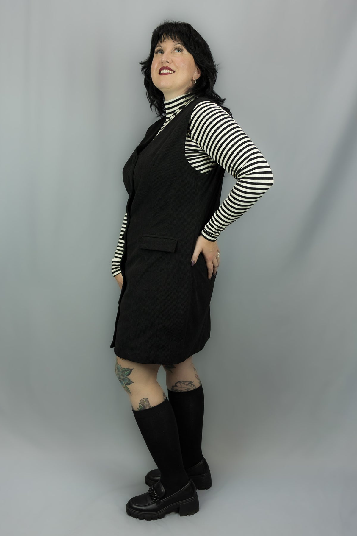 A person modeling a black dress over a black-and-white striped long-sleeve top, paired with black knee-high socks and black shoes, standing against a light gray studio backdrop.