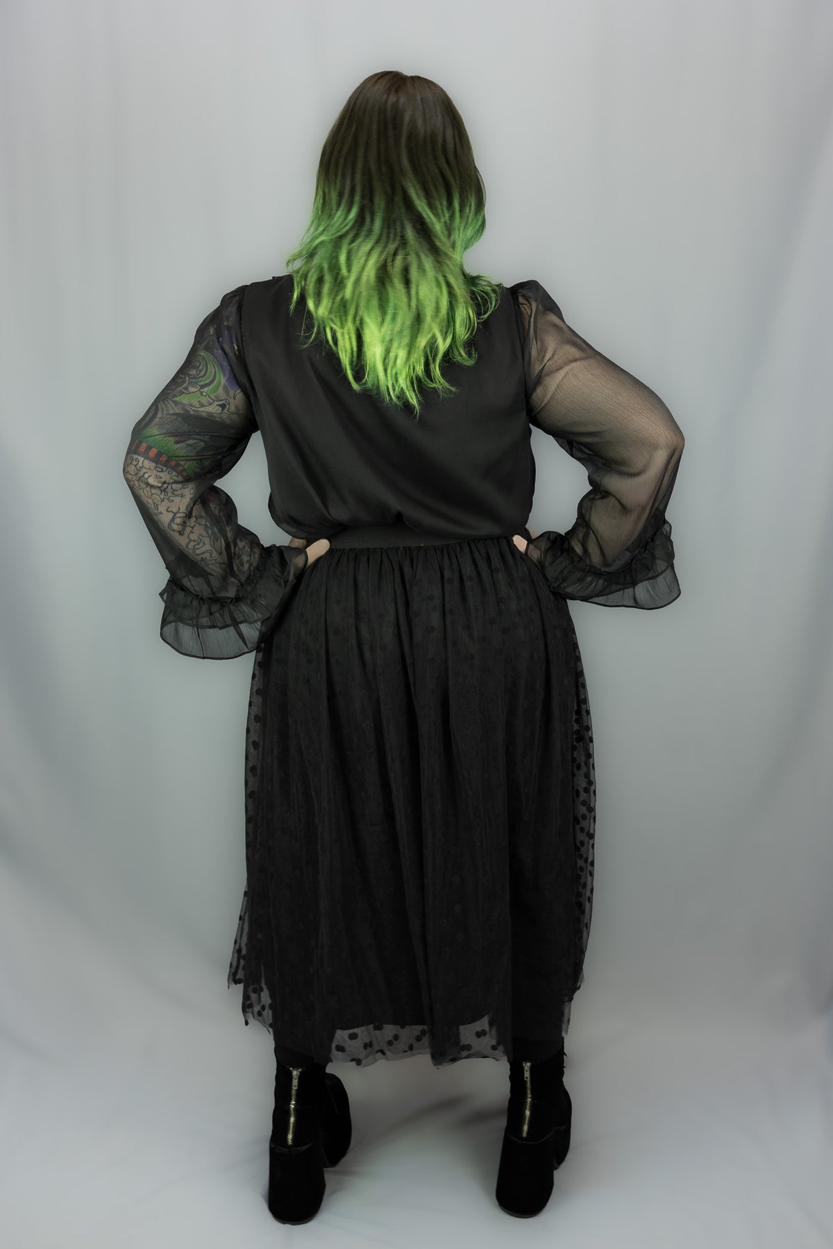 A person shown from the back wearing a black gothic-style outfit with sheer long sleeves, a polka dot skirt, and black platform boots, standing against a light gray studio backdrop.