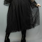 A person wearing a black sheer tulle midi skirt with a subtle polka dot pattern, paired with a black long-sleeve blouse and black platform boots, shown from the back against a light gray studio background.