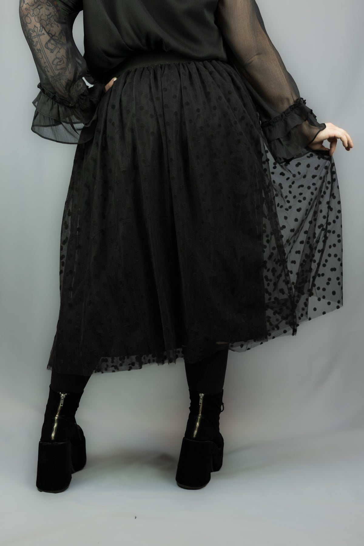 A person wearing a black sheer tulle midi skirt with a subtle polka dot pattern, paired with a black long-sleeve blouse and black platform boots, shown from the back against a light gray studio background.
