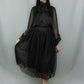 A woman modeling a black gothic-style outfit with sheer long sleeves, a high ruffled collar, and a layered skirt featuring a black polka-dot tulle midi skirt, photographed against a light gray studio backdrop.
