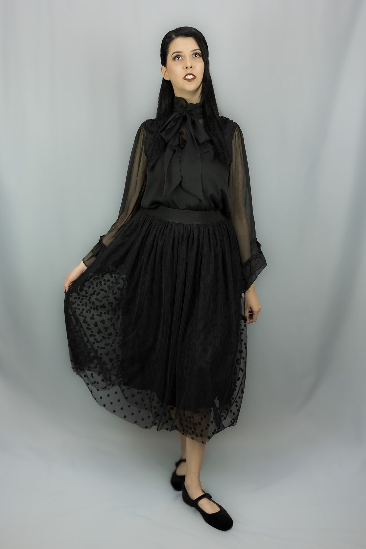 A woman modeling a black gothic-style outfit with sheer long sleeves, a high ruffled collar, and a layered skirt featuring a black polka-dot tulle midi skirt, photographed against a light gray studio backdrop.
