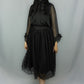 A woman modeling a black midi skirt with sheer long sleeves, a large bow at the neckline, and a gathered skirt with a sheer polka-dot hem, photographed against a light gray studio backdrop.