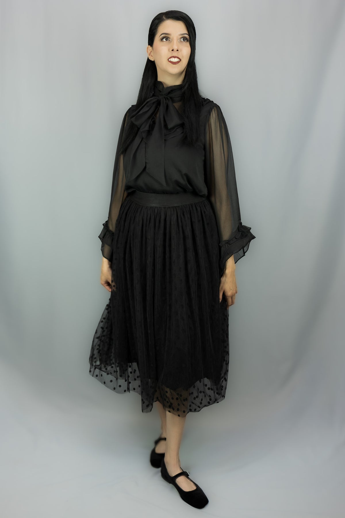 A woman modeling a black midi skirt with sheer long sleeves, a large bow at the neckline, and a gathered skirt with a sheer polka-dot hem, photographed against a light gray studio backdrop.