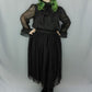 A person modeling a black gothic-style outfit with sheer long sleeves, a large bow at the neckline, and a layered tulle skirt, paired with black lace-up boots, against a light gray studio backdrop.