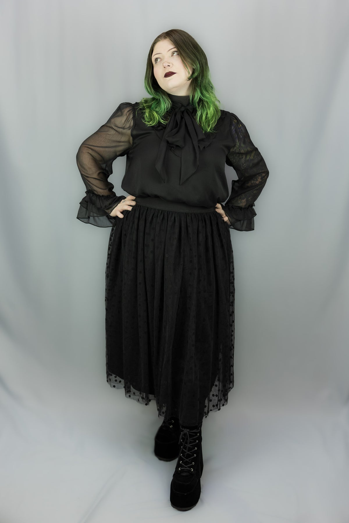 A person modeling a black gothic-style outfit with sheer long sleeves, a large bow at the neckline, and a layered tulle skirt, paired with black lace-up boots, against a light gray studio backdrop.