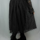 A person wearing a long black sheer tulle midi skirt with a black polka dot pattern, paired with black platform lace-up boots and a black top, photographed against a light gray studio backdrop.