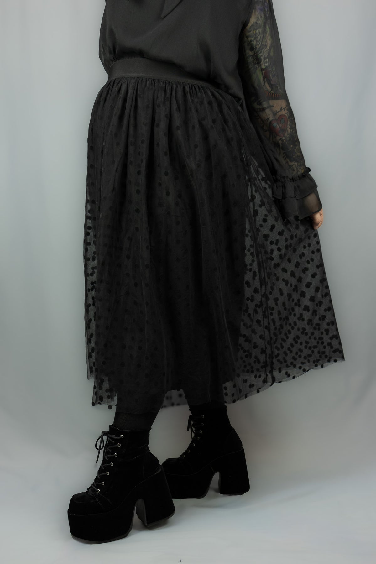 A person wearing a long black sheer tulle midi skirt with a black polka dot pattern, paired with black platform lace-up boots and a black top, photographed against a light gray studio backdrop.