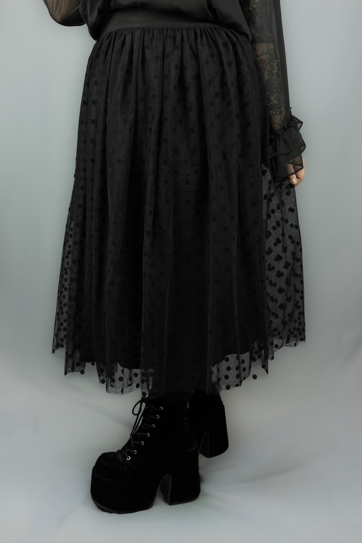 A black midi skirt with a sheer polka-dot overlay and a gathered waist, shown on a model wearing black platform lace-up boots against a light gray background.