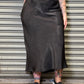 Person wearing a black satin skirt and black t-shirt with a graphic design, standing against a metallic shutter.