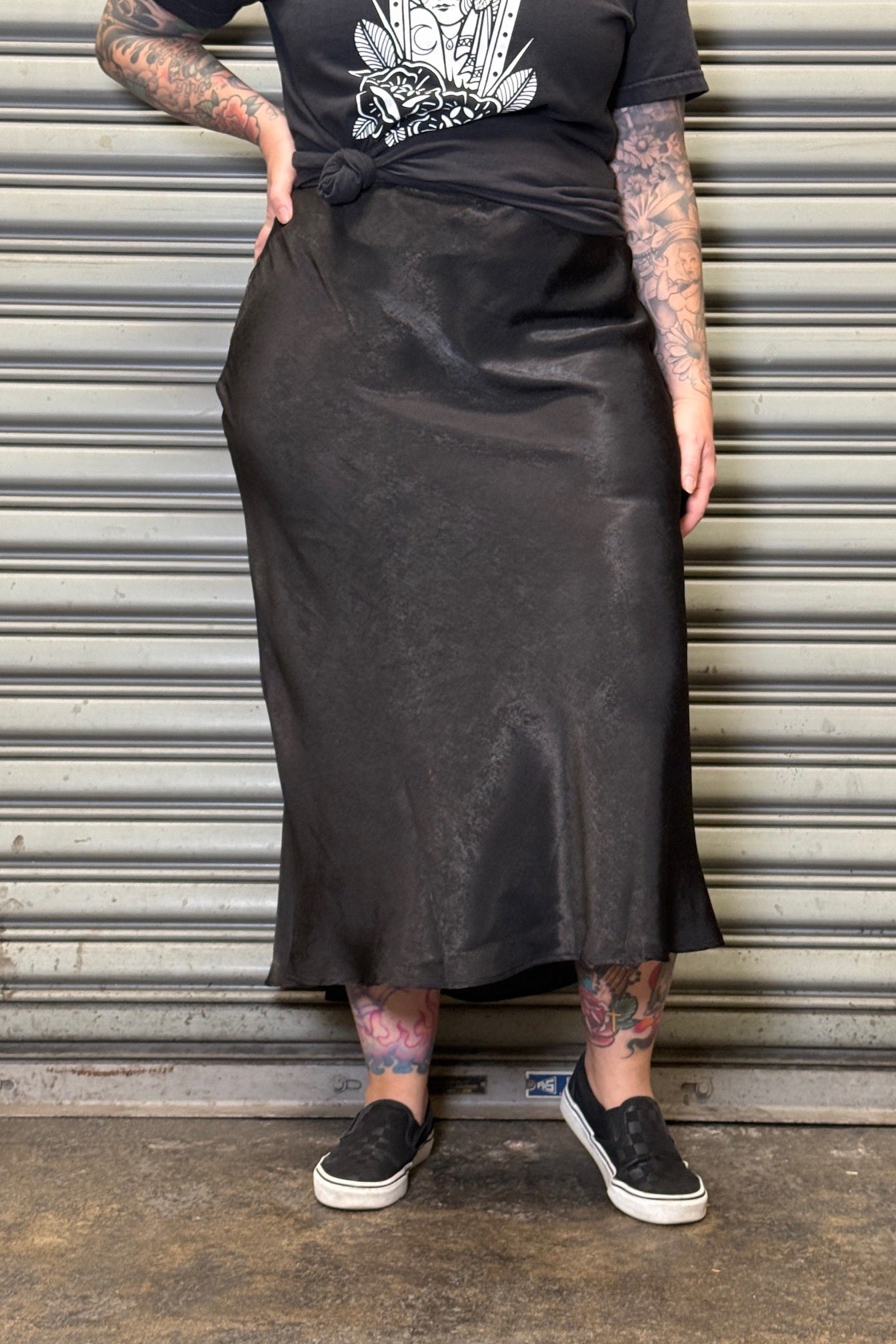 Person wearing a black satin skirt and black t-shirt with a graphic design, standing against a metallic shutter.