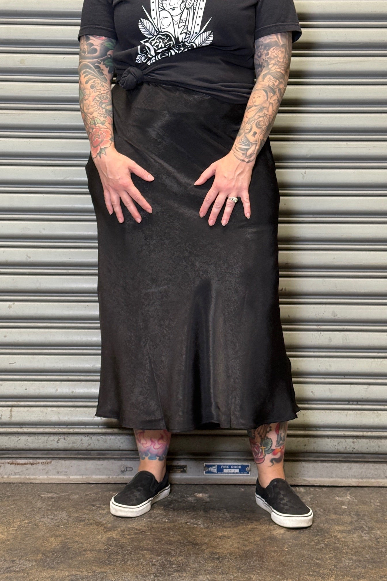 Person wearing a black skirt and black t-shirt with white graphics, standing against a metallic shutter.
