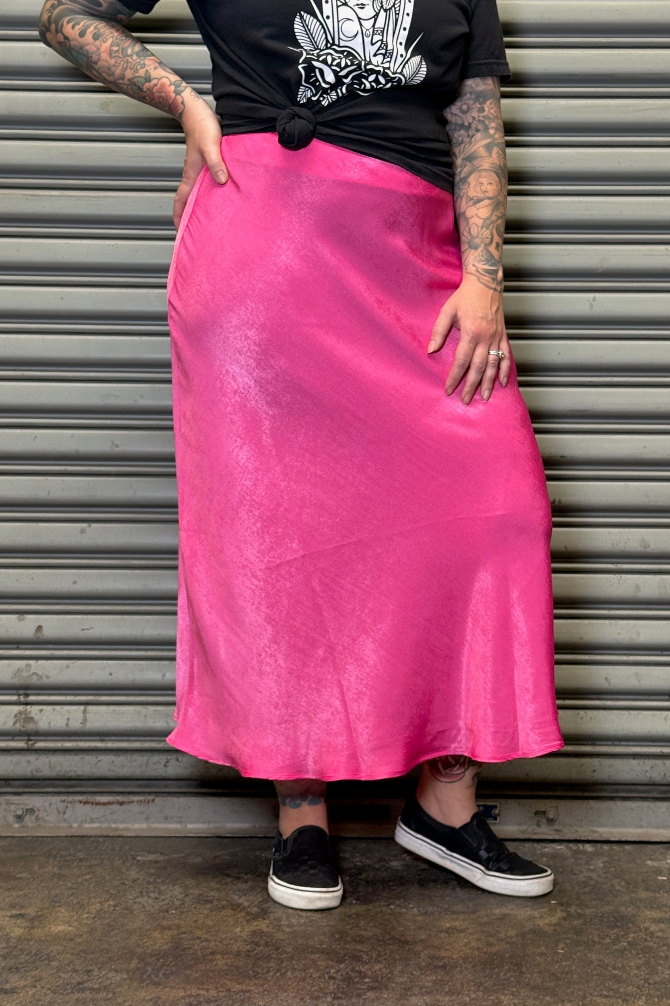 Person wearing a black t-shirt and pink skirt standing against a metal shutter.