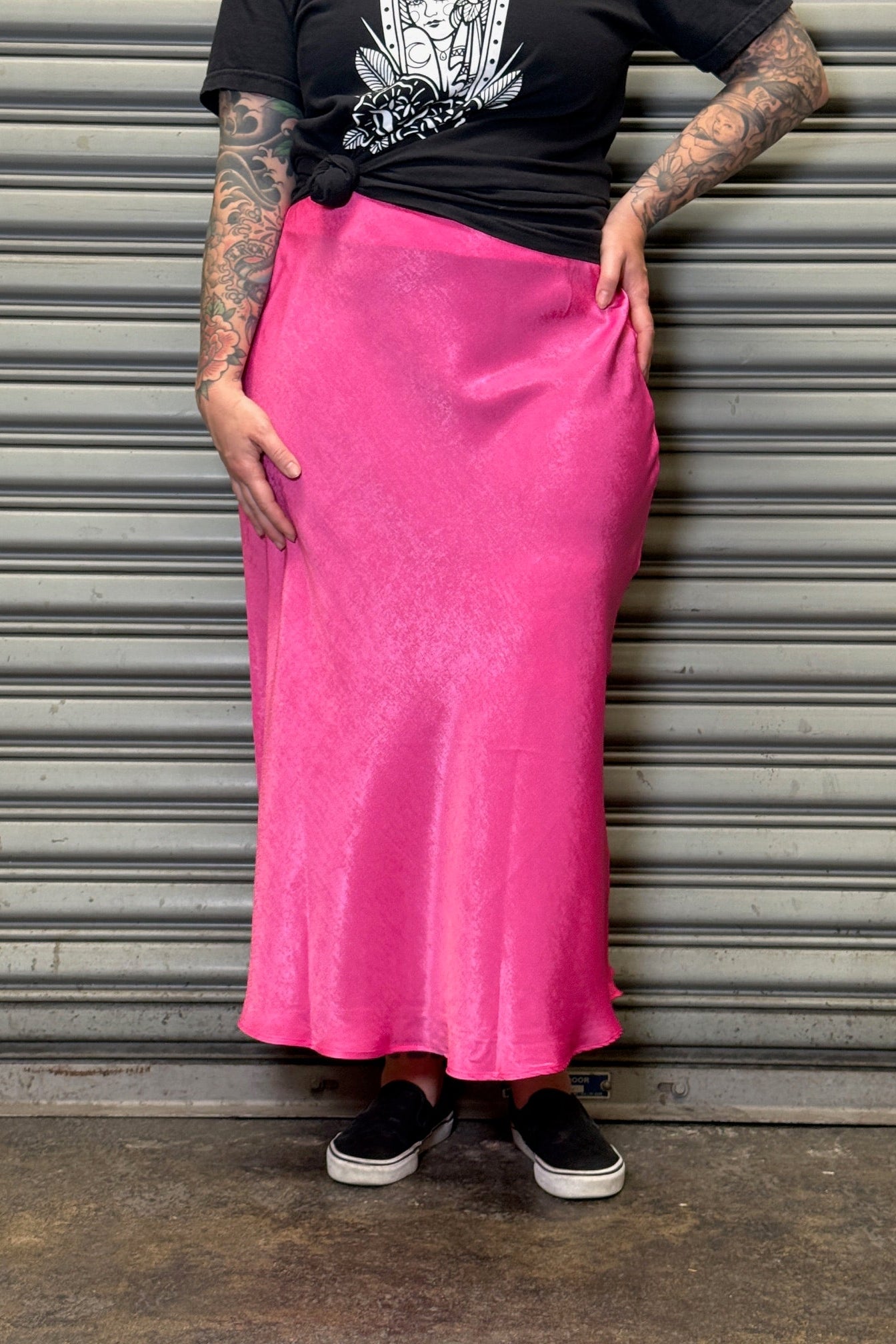 Person wearing a black t-shirt and pink skirt standing against a metallic shutter.
