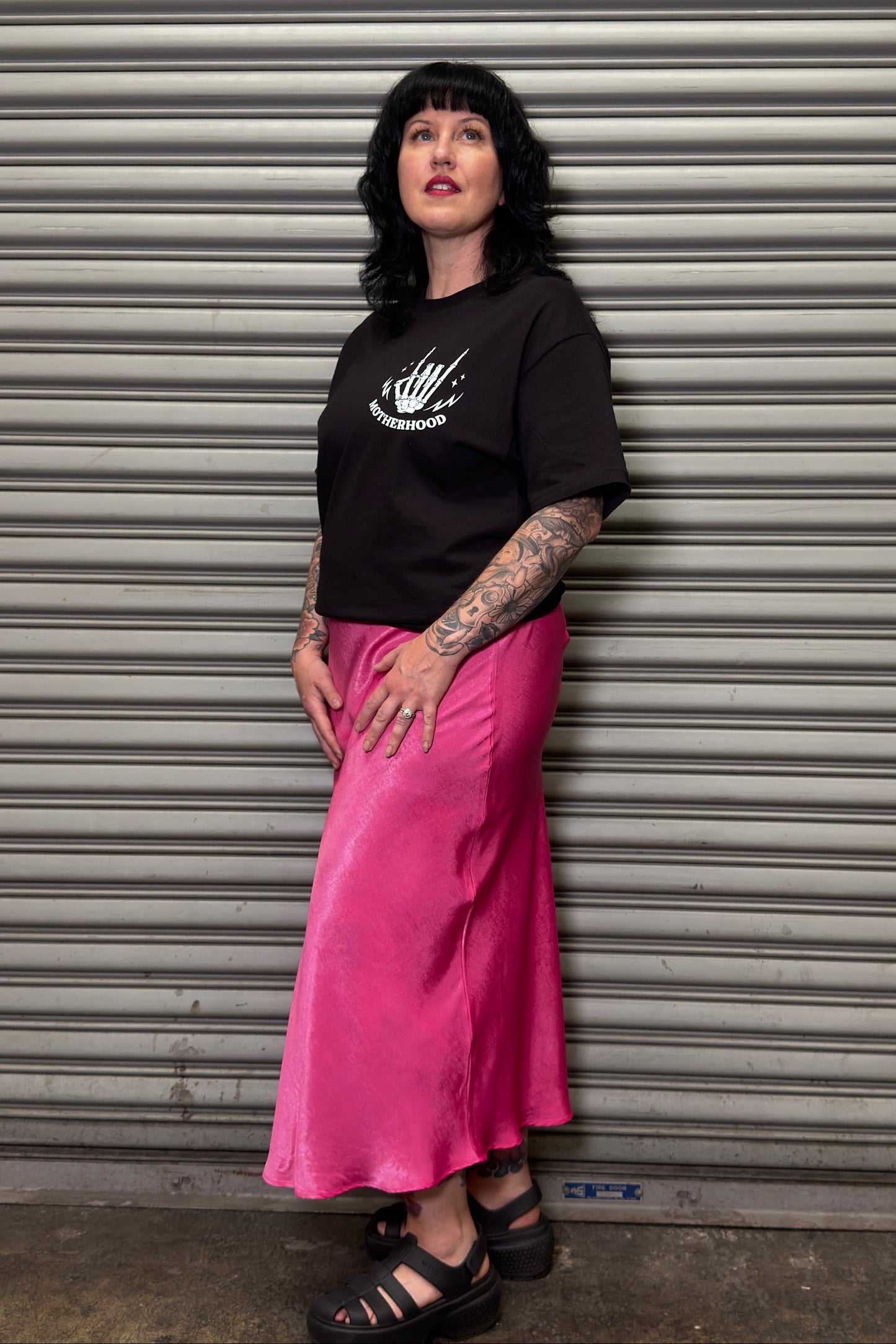 Person wearing a black t-shirt and pink skirt standing in front of a metal shutter.