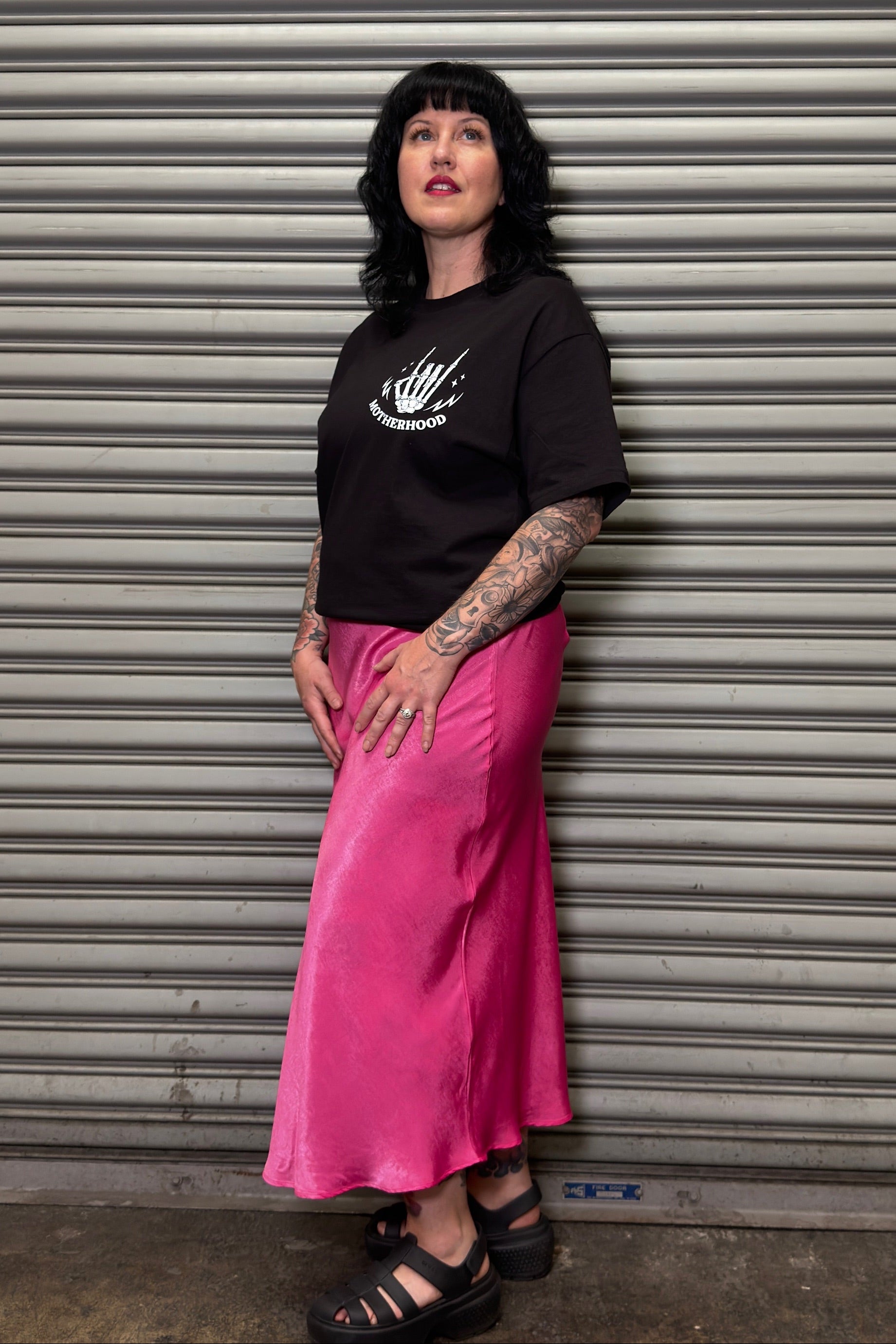 Person wearing a black t-shirt and pink skirt standing in front of a metal shutter.