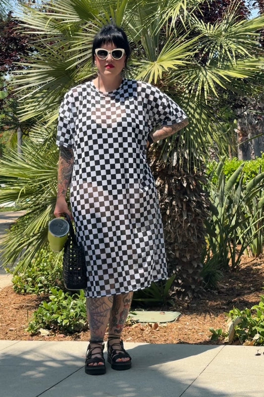 Person wearing a black and white checkered dress standing outdoors with greenery in the background