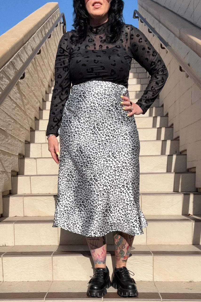 Person wearing a black sheer top and white patterned skirt standing on steps outdoors.