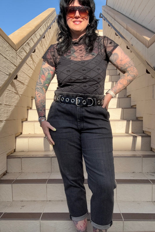 A person wearing a black mesh top with short sleeves and a mock turtle neck, featuring soft velvet bows, paired with black pants.