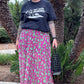 Person wearing a black t-shirt with text and graphics, a pink patterned skirt, and black sandals, standing in front of palm trees.
