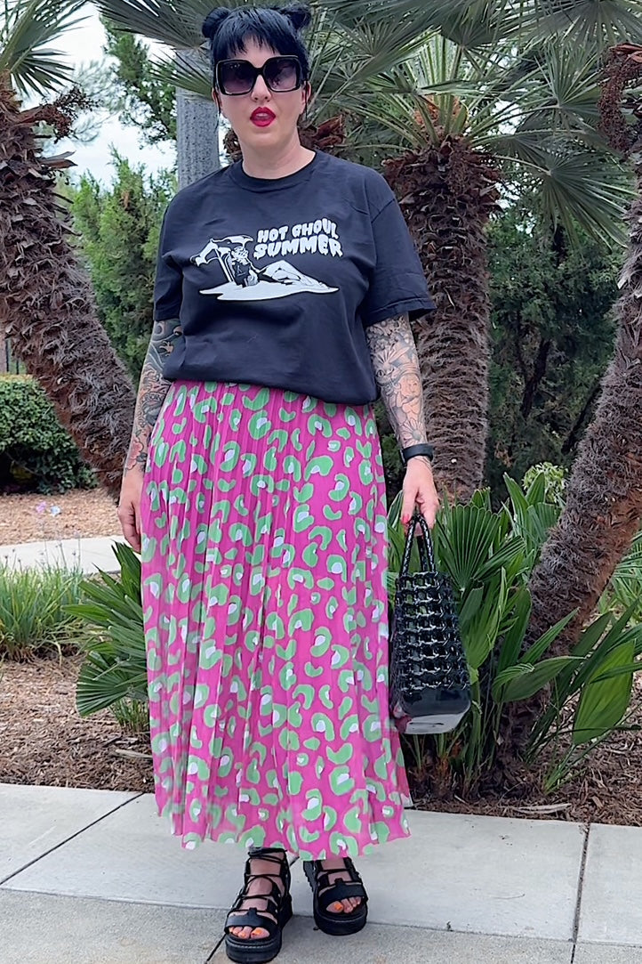 Person wearing a black t-shirt with text and graphics, a pink patterned skirt, and black sandals, standing in front of palm trees.