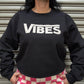 Person wearing a black sweatshirt with 'VIBES' text and pink checkered pants against a metallic shutter background.