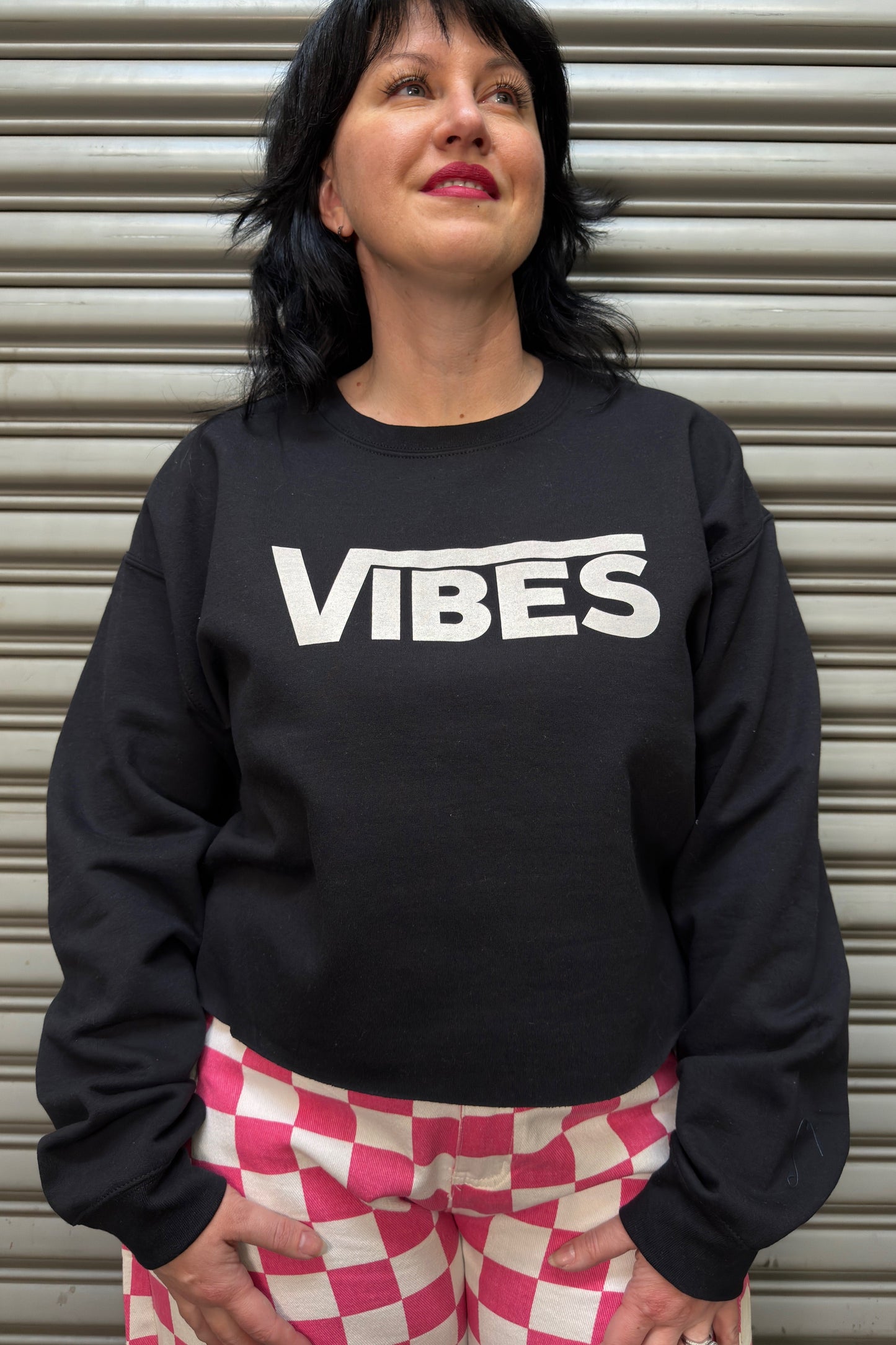 Person wearing a black sweatshirt with 'VIBES' text in front of a metallic shutter.