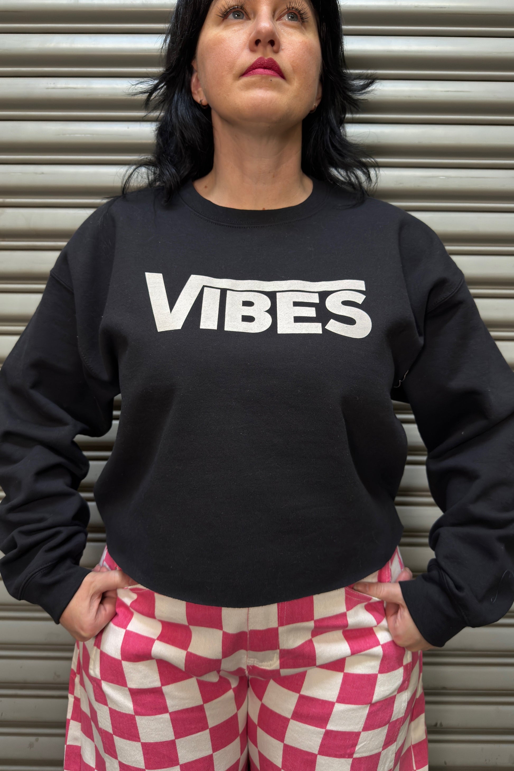 Person wearing a black sweatshirt with 'VIBES' text and pink checkered pants against a metallic shutter background.