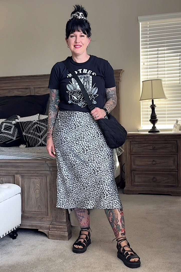 A person wearing a black top with a graphic design and an A-line midi skirt with a leopard print pattern. They are also wearing black strappy sandals and have a bag and a watch visible.