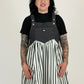 Person wearing a black denim pinafore over a black turtleneck and white striped dress on a plain background