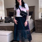 A person wearing a midi-length pleated skirt with iridescent turquoise and purple colors, paired with a white top and pink hat, standing in a room with a bed and sunglasses on a surface in the background.