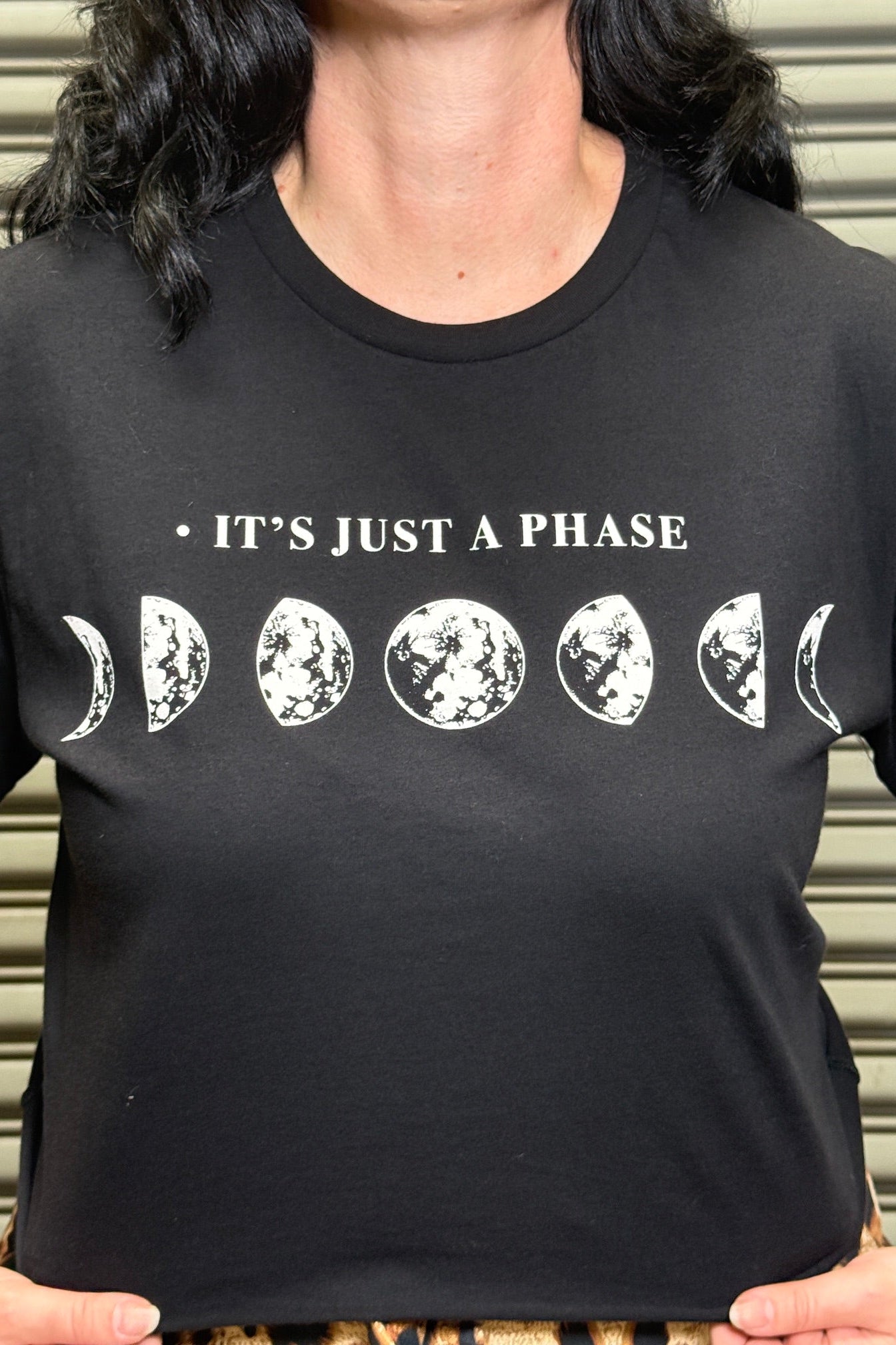 Black t-shirt with moon phase design and 'It's Just a Phase' text worn by a person.