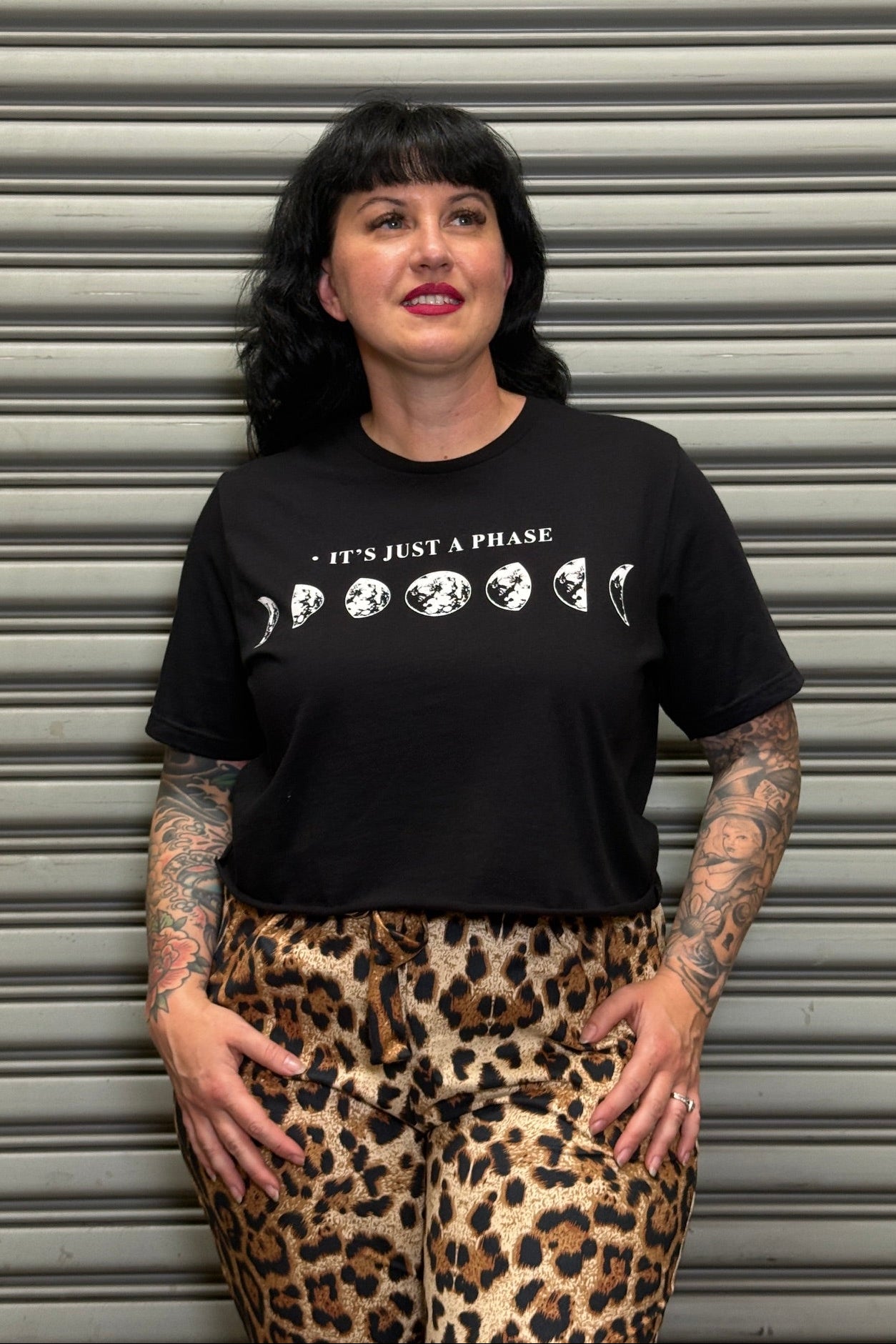 Person wearing a black t-shirt with white text and graphics, standing in front of a metallic shutter.