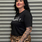 Person wearing a black t-shirt with white text and leopard print pants standing in front of a gray shutter.