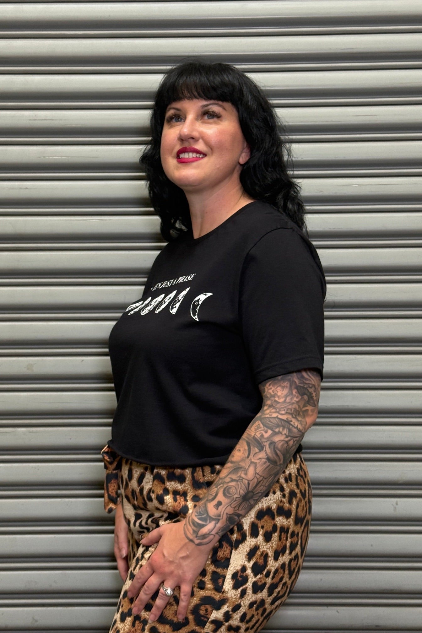 Person wearing a black t-shirt with white text and leopard print pants standing in front of a gray shutter.