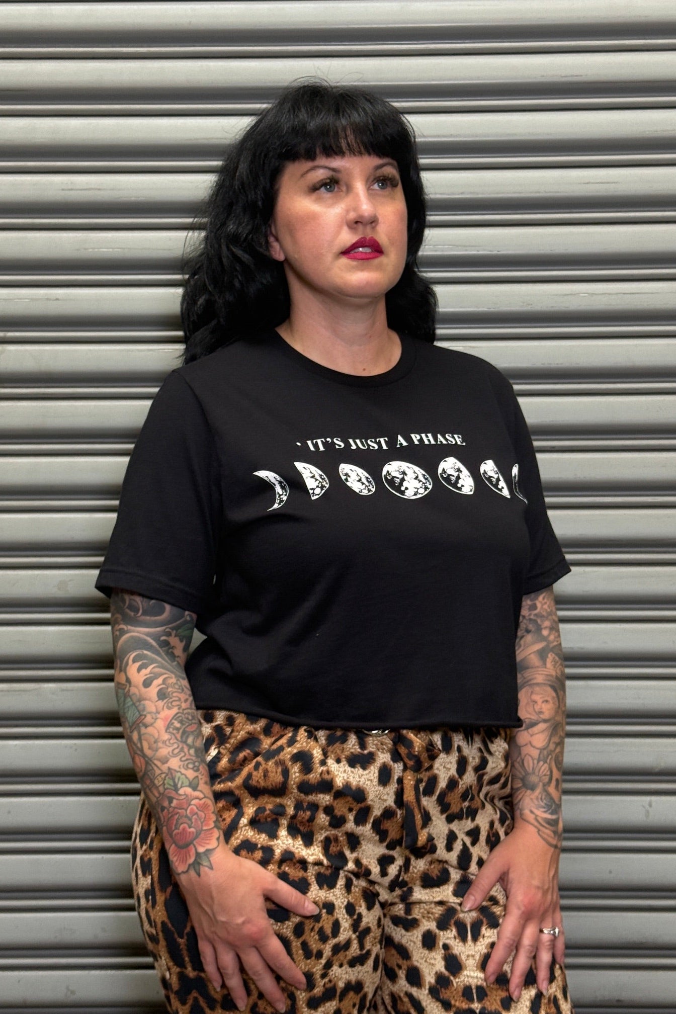 Person wearing a black t-shirt with text and moon phases graphic, standing in front of a metallic shutter.