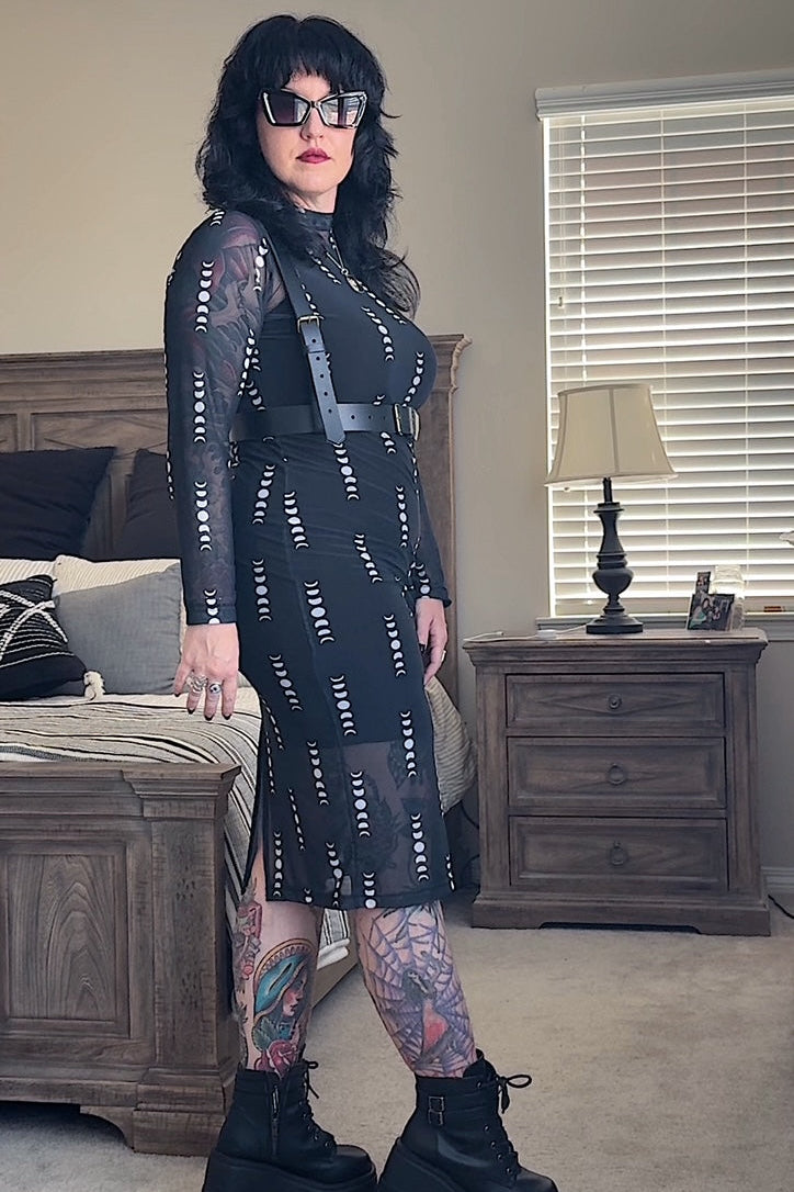 Person wearing a black patterned dress and black boots in a bedroom setting.