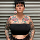 Woman with black hair and tattoos wearing a black camisole and plaid high waist pants standing in front of a metal back drop.