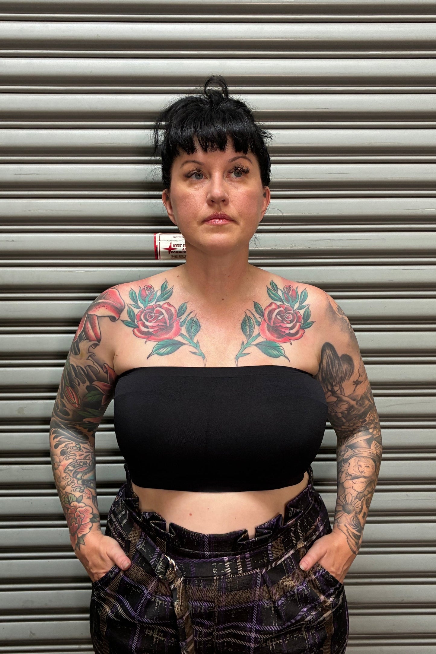Woman with black hair and tattoos wearing a black camisole and plaid high waist pants standing in front of a metal back drop.