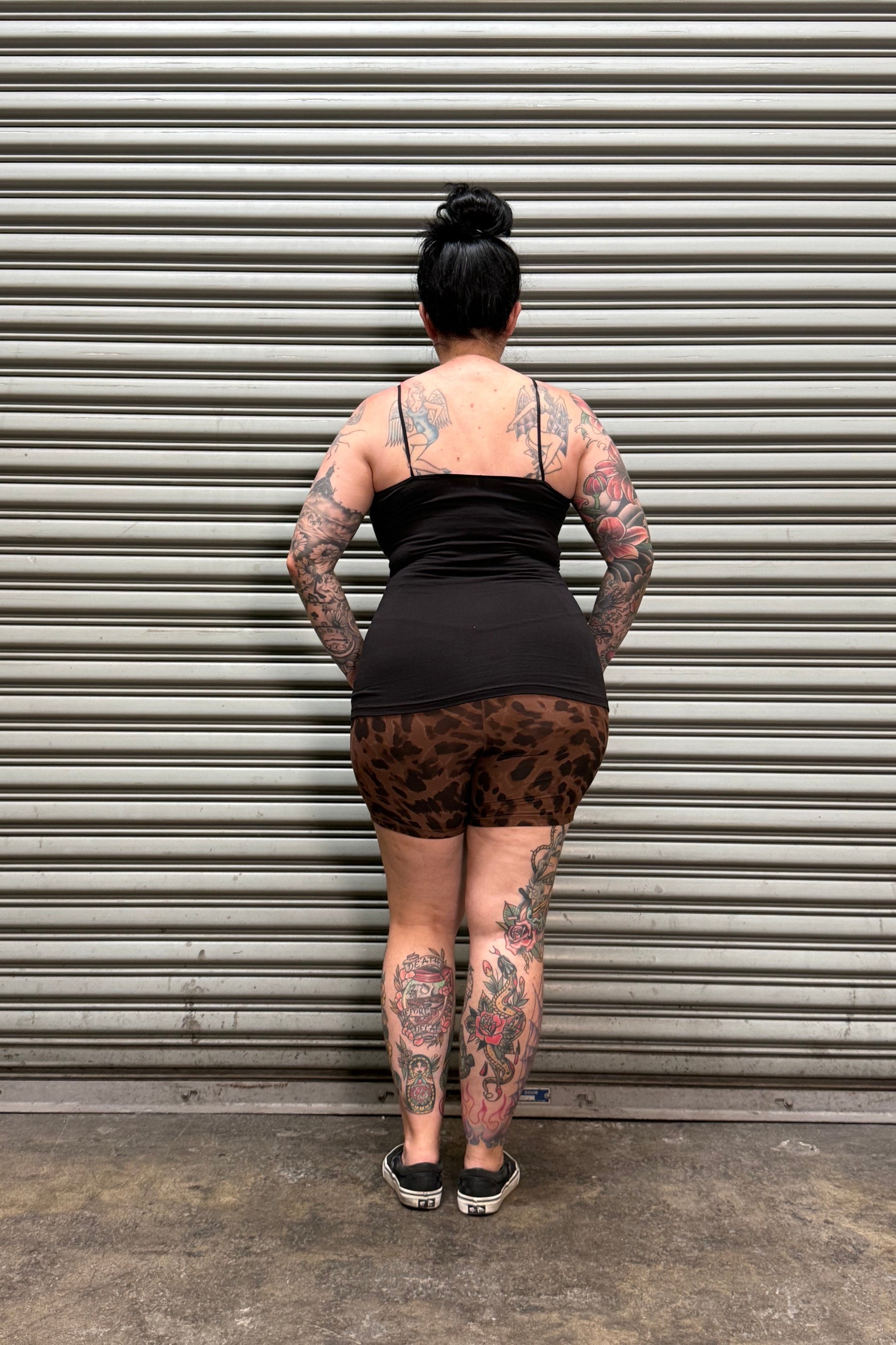 A person viewed from above, wearing a black sleeveless top and leopard-print shorts, standing against a gray corrugated metal wall.