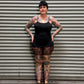 A tattooed woman wearing a black sleeveless top and leopard-print leggings stands with hands on hips in front of a gray metal roll-up door.