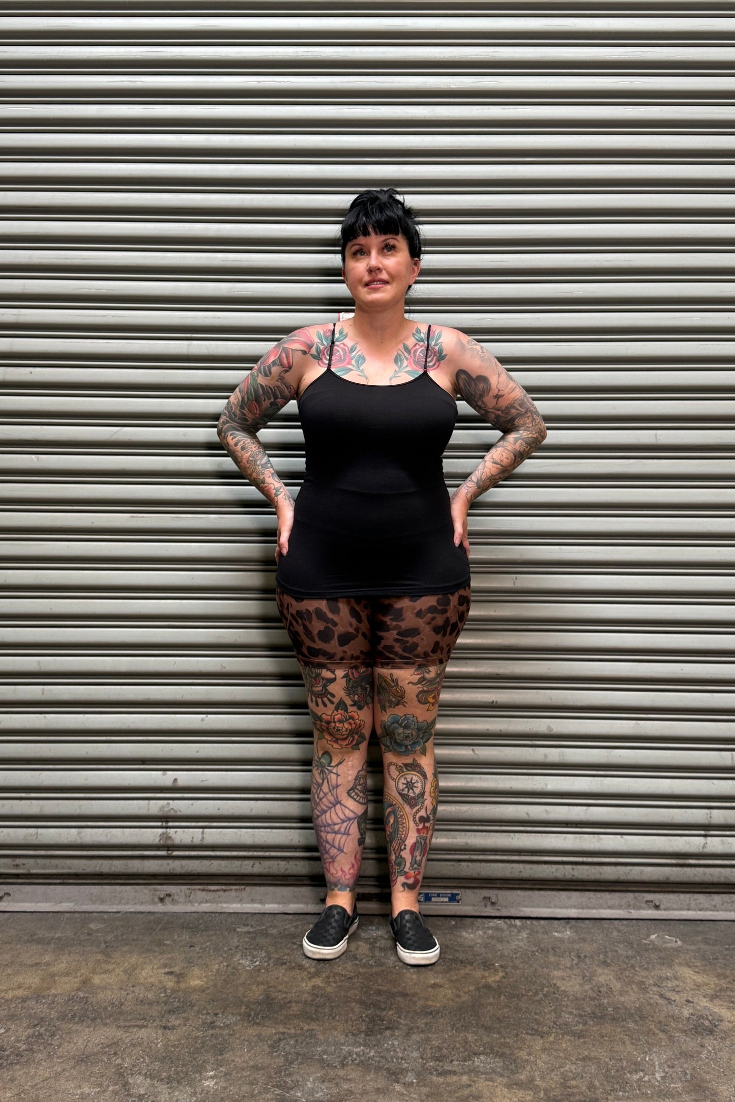 A tattooed woman wearing a black sleeveless top and leopard-print leggings stands with hands on hips in front of a gray metal roll-up door.