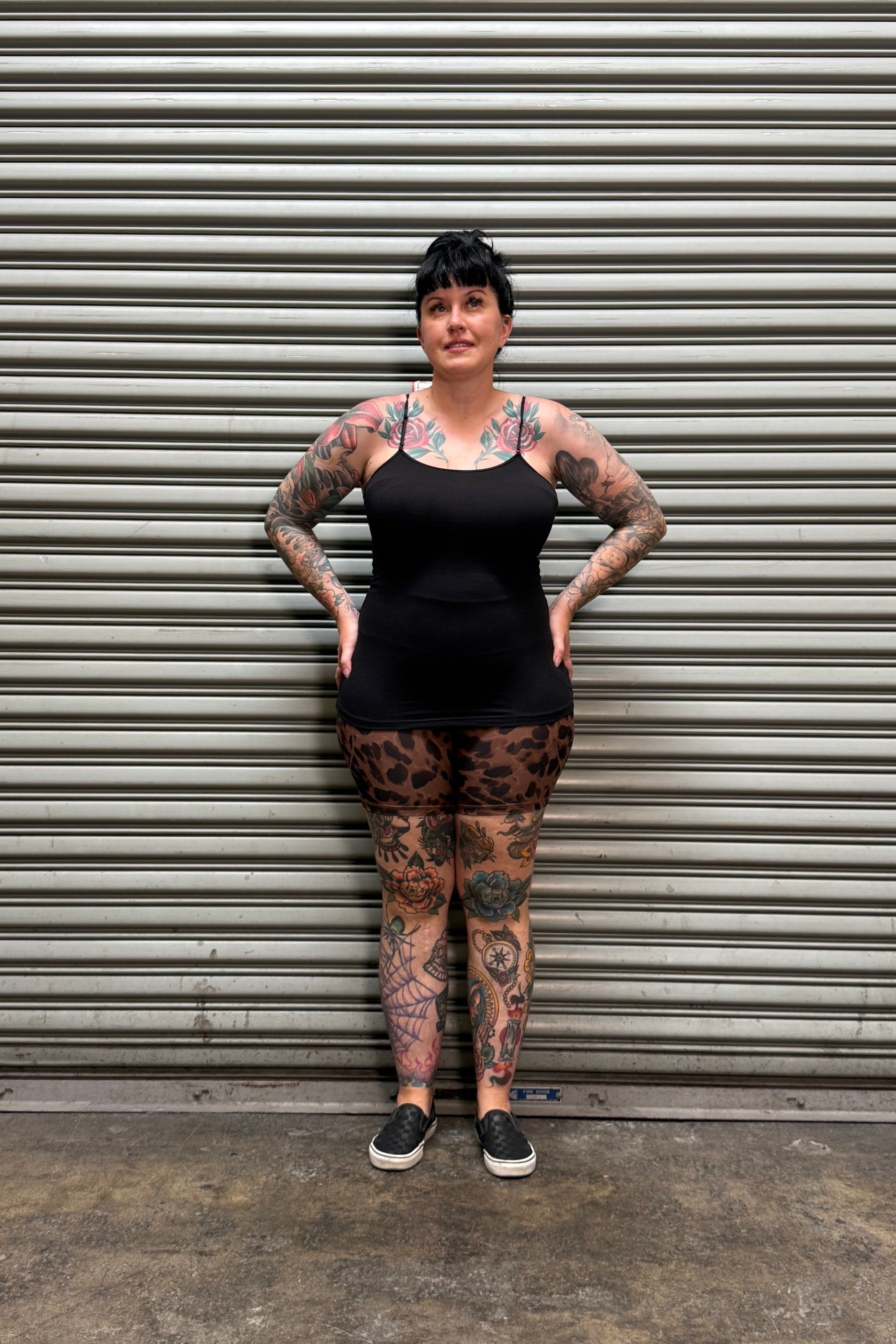 A tattooed woman wearing a black sleeveless top and leopard-print leggings stands with hands on hips in front of a gray metal roll-up door.