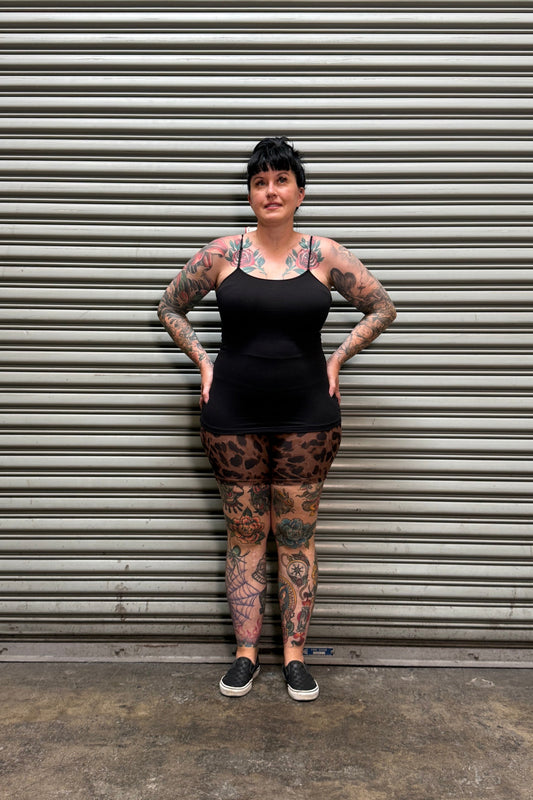 A tattooed woman wearing a black sleeveless top and leopard-print leggings stands with hands on hips in front of a gray metal roll-up door.