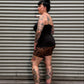 A tattooed woman wearing a black tank top and leopard-print shorts leans against a gray corrugated metal wall next to a concrete floor.
