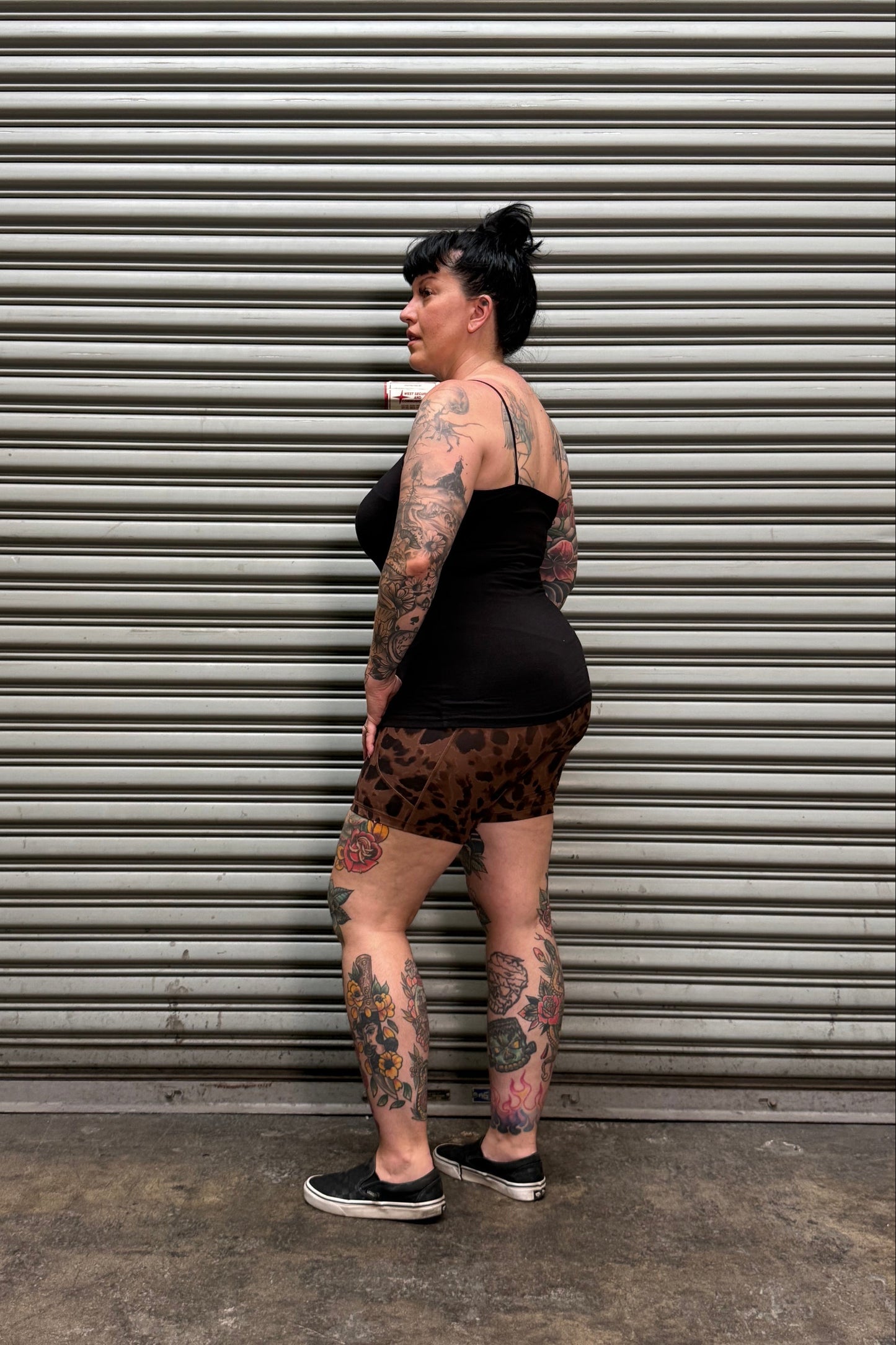 A tattooed woman wearing a black tank top and leopard-print shorts leans against a gray corrugated metal wall next to a concrete floor.