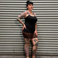 A tattooed woman wearing a black tank top and leopard-print mini skirt poses against a gray corrugated metal wall.
