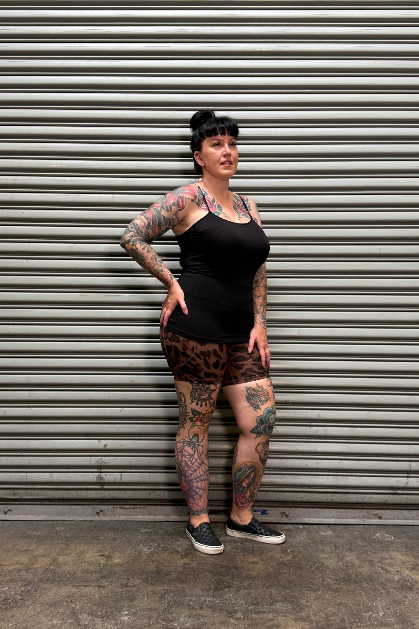 A tattooed woman wearing a black tank top and leopard-print mini skirt poses against a gray corrugated metal wall.