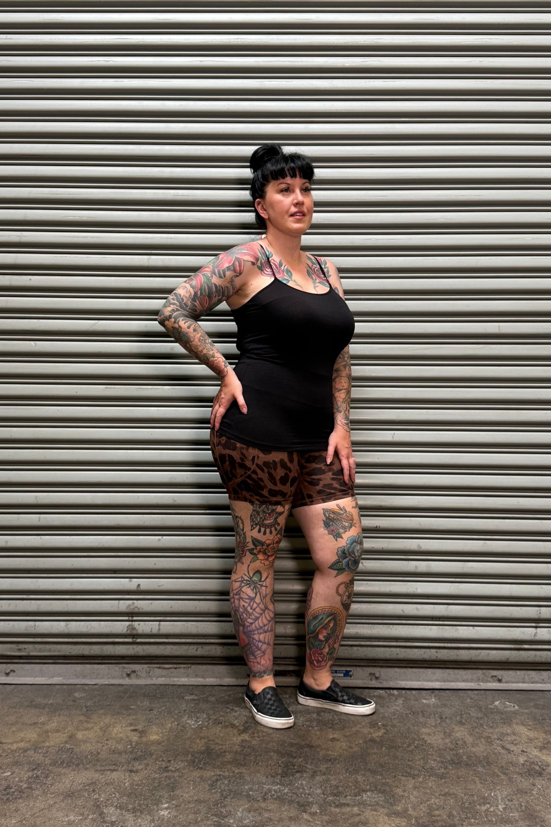 A tattooed woman wearing a black tank top and leopard-print mini skirt poses against a gray corrugated metal wall.