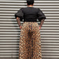 A person viewed from above wearing a black top and a leopard-print skirt, standing in front of a corrugated metal wall.